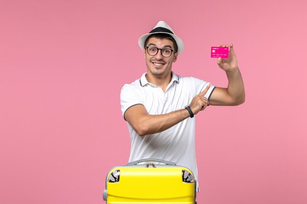 Front view of young man holding bank card on summer vacation on a light-pink wall