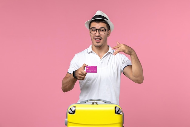 Front view of young man holding bank card on summer vacation on light-pink wall