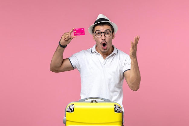 Front view of young man holding bank card on summer vacation on light-pink wall