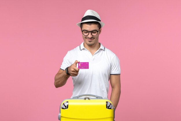 Front view of young man holding bank card on summer vacation on pink wall