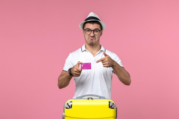 Front view of young man holding bank card on summer vacation on pink wall