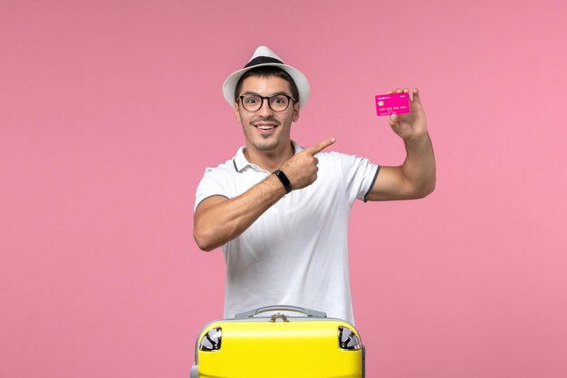 Front view of young man holding bank card on vacation on light-pink wall