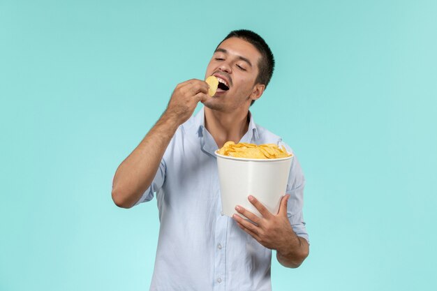 Front view young man holding basket with cips and eating on blue wall film remote movie cinema male