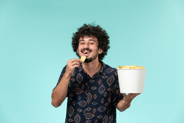 Front view young man holding basket with potato cips and eating on a light blue wall cinema movie film theater male