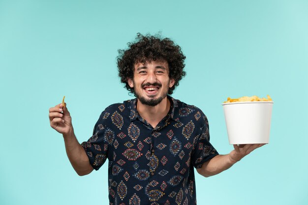 Front view young man holding basket with potato cips and eating them on a blue wall cinema movie film theater male