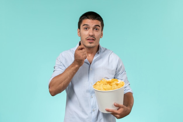 Front view young man holding basket with potato cips eating and watching movie on a light blue wall lonely remote movie cinema