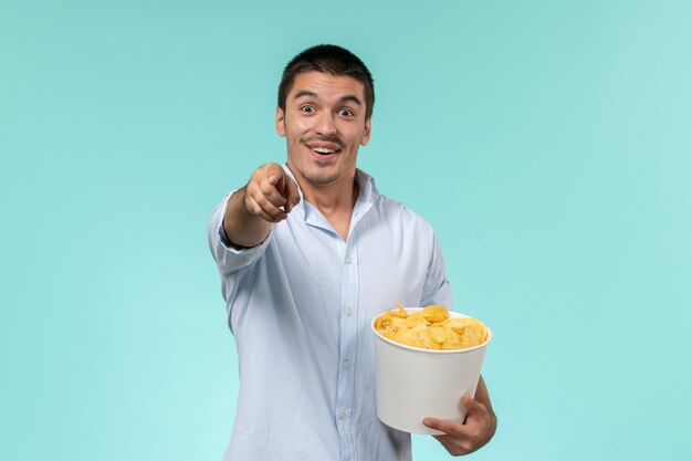 Вид спереди молодой человек, держащий корзину с картофельными чипсами, ест и смотрит фильм на голубой стене одинокий удаленный кинотеатр
