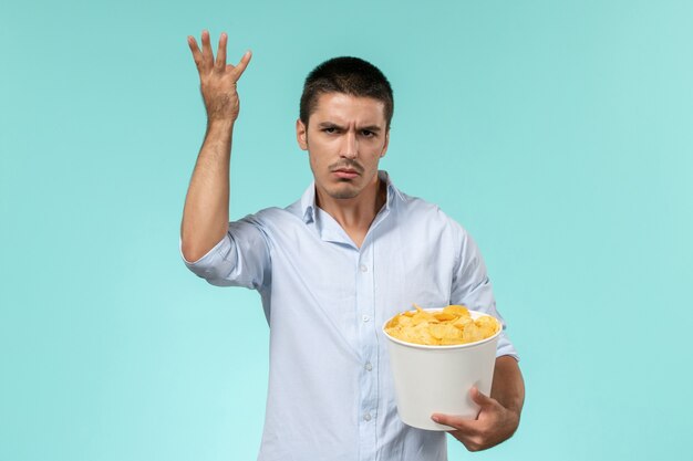 Вид спереди молодой человек, держащий корзину с картофельными чипсами, ест и смотрит фильм на голубой стене одинокий удаленный кинотеатр