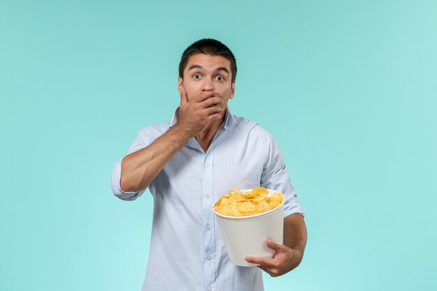 Front view young man holding basket with potato cips eating and watching movie shocked on blue wall lonely remote movies cinema