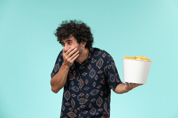 Front view young man holding basket with potato cips and laughing on blue wall remote cinema film movies theater