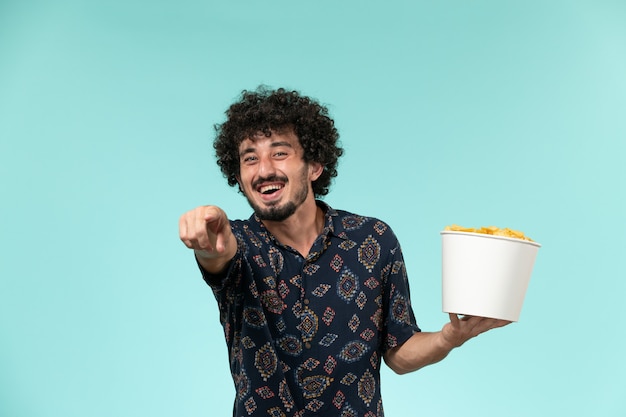 Front view young man holding basket with potato cips and laughing out loud on blue wall remote cinema film movies theater