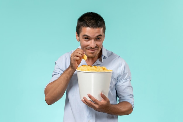 Front view young man holding basket with potato cips on light-blue wall film remote movie cinema