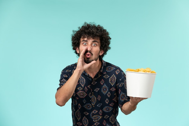 Вид спереди молодой человек, держащий корзину с картофельными чипсами на голубой стене удаленного кинотеатра, кинотеатра