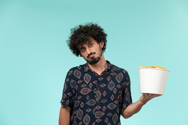 Front view young man holding basket with potato cips on light-blue wall remote film cinema movie theater