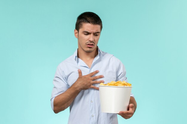 Front view young man holding basket with potato cips on a light blue wall remote movie cinema lonely male