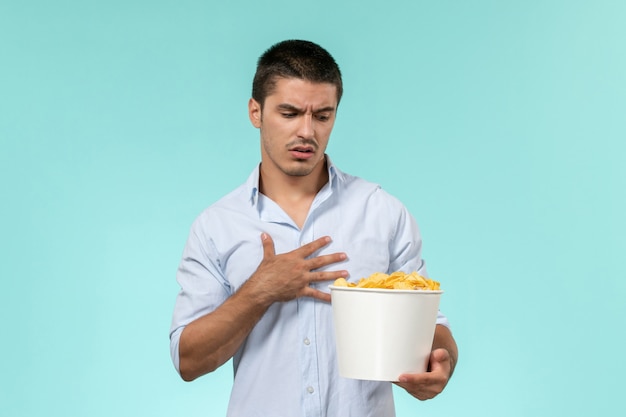 Front view young man holding basket with potato cips on a light blue wall remote movie cinema lonely male