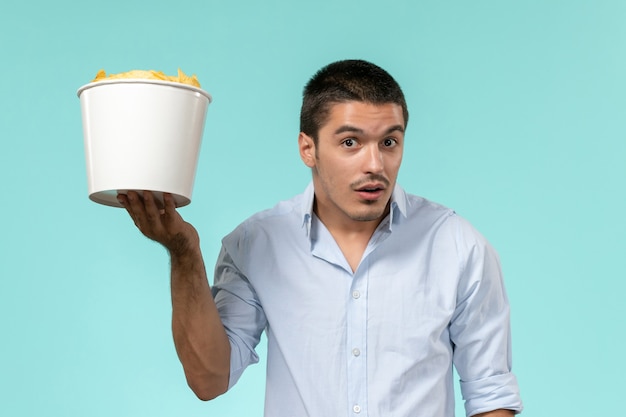 Front view young man holding basket with potato cips on the light-blue wall remote movie cinema lonely male