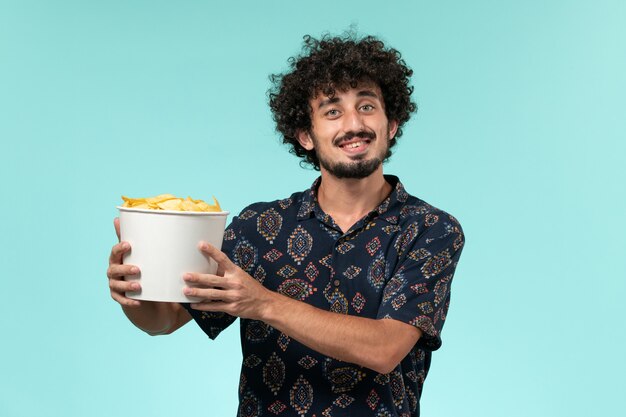 Front view young man holding basket with potato cips and watching movie on blue wall film cinema movie male remote