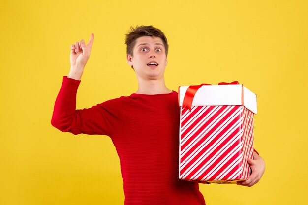 黄色の壁に大きなクリスマスプレゼントを保持している若い男の正面図