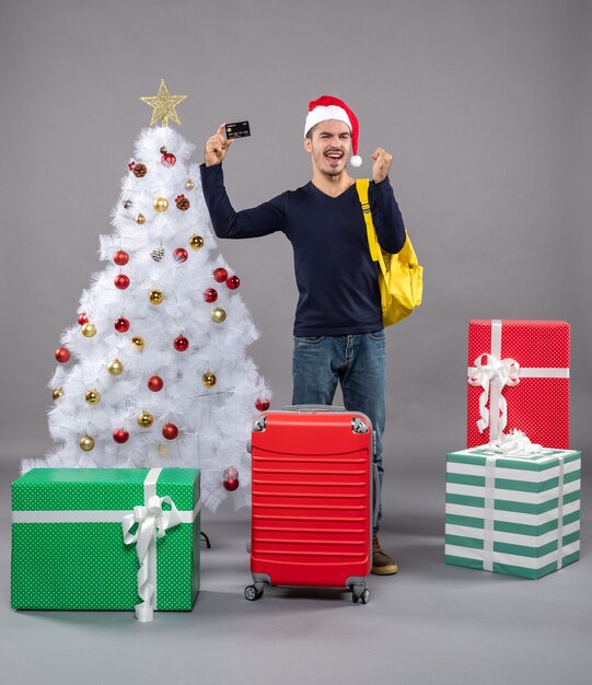 Front view young man holding card standing near xmas tree and presents on grey isolated