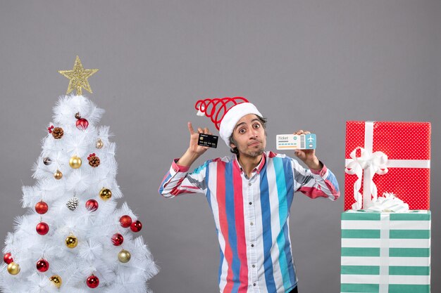 Front view young man holding card and travel ticket near xmas tree with colorful xmas toys