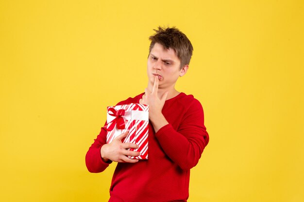 Front view of young man holding christmas present confused on yellow wall