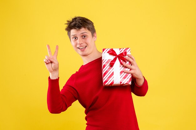 Front view of young man holding christmas present onsing yellow wall