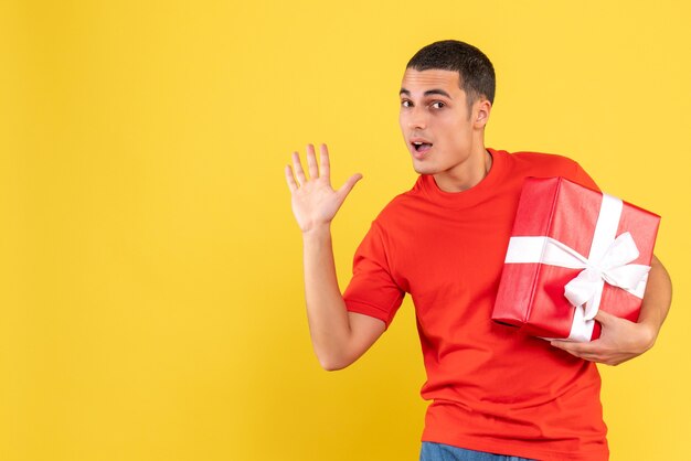 Front view of young man holding christmas present waving on yellow wall