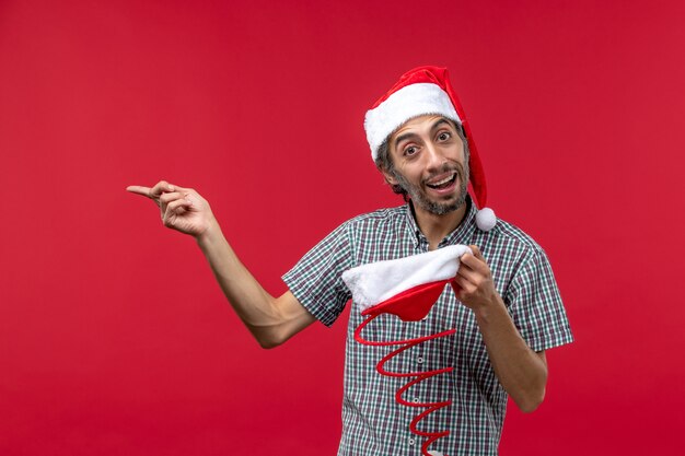 Вид спереди молодого человека, держащего шапку рождественской игрушки на светло-красной стене