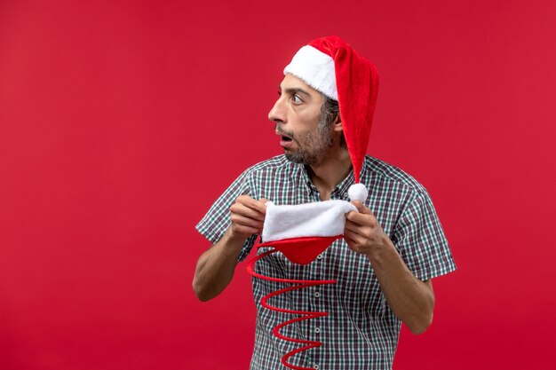 Вид спереди молодого человека, держащего шапку рождественской игрушки на красной стене