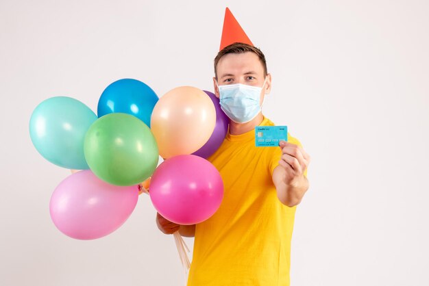 Front view of young man holding colorful balloons and bank card in mask on white wall
