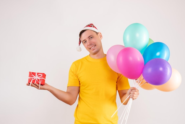 Front view of young man holding colorful balloons and little present on white wall