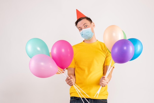 Front view of young man holding colorful balloons in mask on a white wall