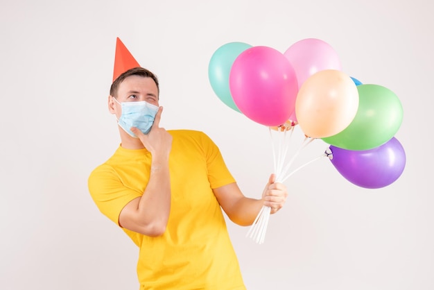 Front view of young man holding colorful balloons in mask on a white wall