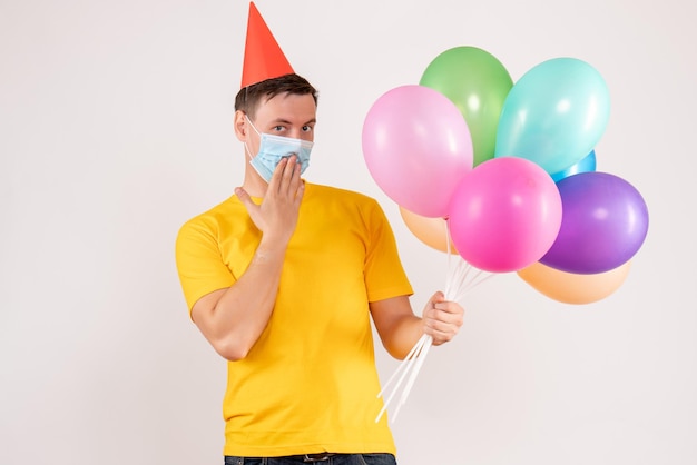 Front view of young man holding colorful balloons in mask on a white wall