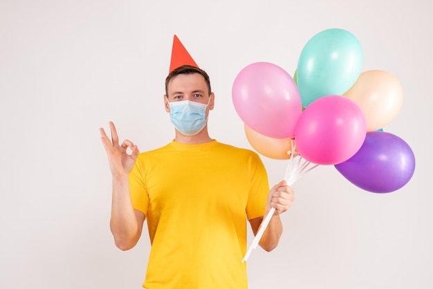 Front view of young man holding colorful balloons in mask on a white wall