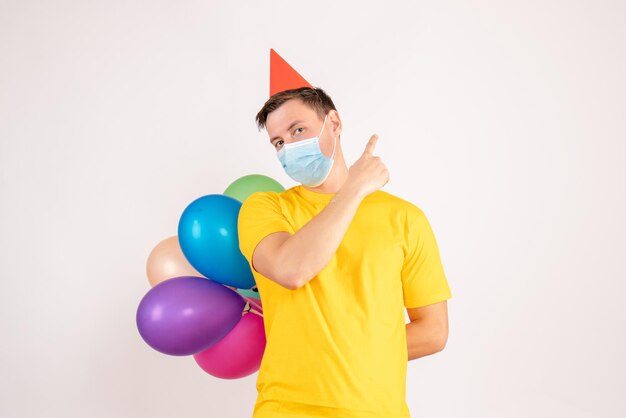 Front view of young man holding colorful balloons in mask on a white wall
