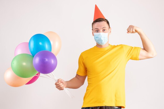 Front view of young man holding colorful balloons in mask on a white wall