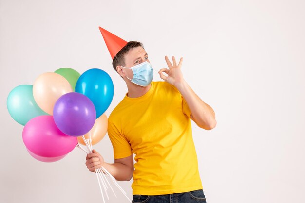 Front view of young man holding colorful balloons in mask on a white wall