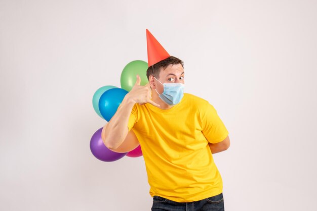 Front view of young man holding colorful balloons in mask on a white wall