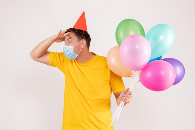 Front view of young man holding colorful balloons in mask on a white wall