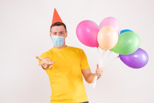 Вид спереди молодого человека, держащего разноцветные шары в маске на белой стене
