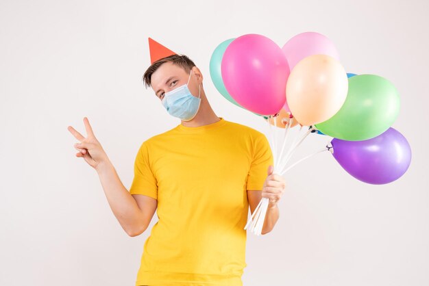 Front view of young man holding colorful balloons in mask on the white wall