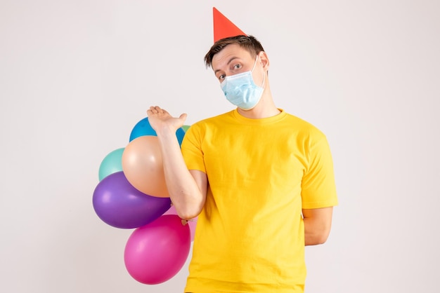 Front view of young man holding colorful balloons in mask on the white wall