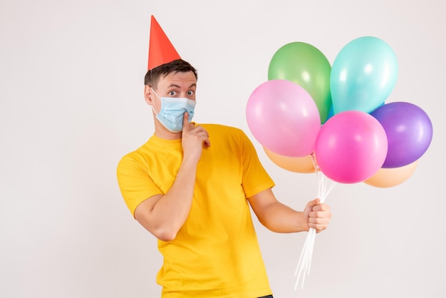 Front view of young man holding colorful balloons in mask on the white wall