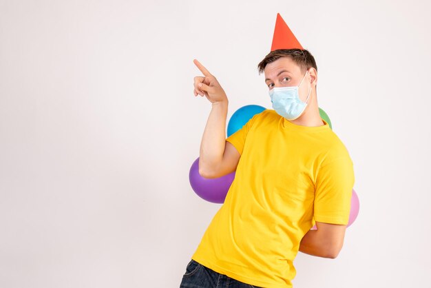 Front view of young man holding colorful balloons in mask on the white wall