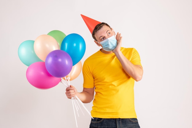 Front view of young man holding colorful balloons in mask on the white wall