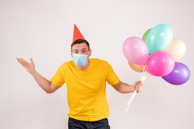 Front view of young man holding colorful balloons in mask on the white wall