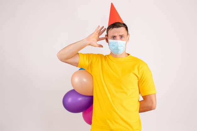 Front view of young man holding colorful balloons in mask on the white wall
