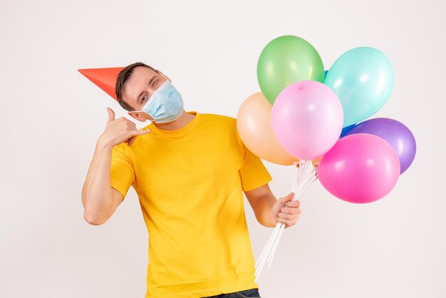Front view of young man holding colorful balloons in mask on the white wall
